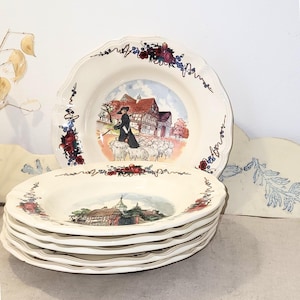 Puede incluir: Una pila de platos de cerámica blanca con bordes festoneados. Cada plato presenta una escena pastoral central y un borde floral en rojo, azul y verde. Los platos tienen una estética vintage.