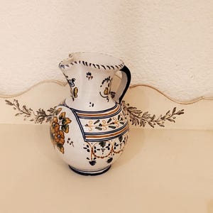 Może przedstawiać: Biały dzbanek ceramiczny z niebieskim i pomarańczowym wzorem kwiatowym. Dzbanek ma uchwyt i dzióbek.