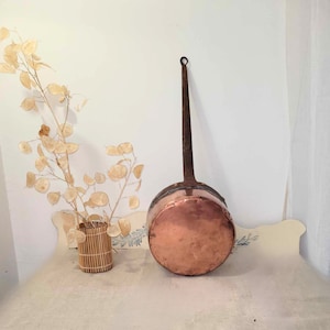 Peut inclure: Une casserole en cuivre vintage avec une longue poignée en fer. La casserole est posée sur une surface blanche.