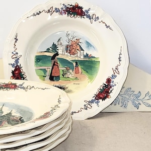 Puede incluir: Platos color crema con bordes festoneados. Cada plato presenta una escena de paisaje central con figuras y una iglesia, enmarcada por diseños florales y de vid en rojo, azul y verde. Los platos tienen una estética vintage.