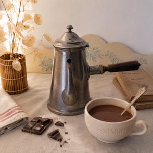以下が含まれることがあります： 木製のハンドルが付いた銀色のコーヒーポットが、ホットチョコレートが入った白いティーカップの隣に置かれています。 近くには、ダークチョコレートバーとスプーンがあります。 竹の花瓶に入ったドライフラワーも存在します。