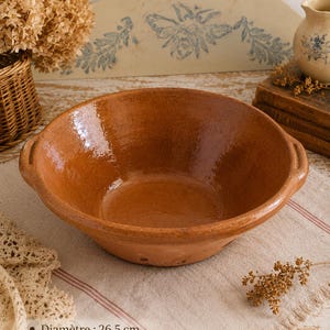 May include: A round, glazed terracotta bowl with two small side handles. The bowl has a warm, reddish-brown finish and a smooth, reflective interior surface. Text printed on the image indicates a diameter of 26.5 cm and a height of 10 cm.