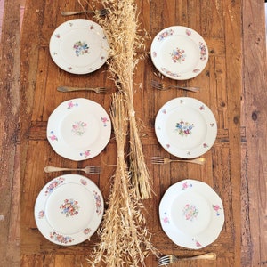 May include: Six white ceramic plates with floral designs on a wooden table with dried wheat stalks in the center.
