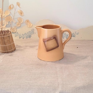 Peut inclure: Pichet en céramique beige avec un motif de biscuit marron sur le devant. Le pichet a une anse et un bec verseur. L'arrière-plan comprend un panier en osier et des plantes séchées.