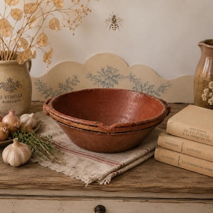 May include: A rustic, round, terracotta-colored earthenware bowl with a wide, open top and small side handles. The bowl features a textured, weathered surface with dark, horizontal linear markings around the exterior rim and a shallow spout.