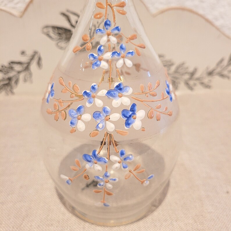K&ouml;nnte beinhalten: Eine klare Glasflasche in Tropfenform mit einem zarten Blumenmuster. Die Flasche ist mit handbemalten blauen und wei&szlig;en Blumen mit braunen Stielen verziert.