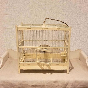 Puede incluir: Una jaula de pájaros blanca con un marco de madera y una jaula de alambre metálico. La jaula tiene una pequeña puerta en la parte delantera y una percha en el interior. La jaula está sentada sobre una superficie blanca.