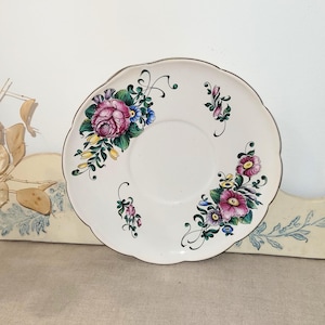 Könnte beinhalten: Eine weiße Keramiksaucerschale mit einem gewellten Rand und einem Blumendesign. Das Design zeigt rosa, gelbe, blaue und grüne Blumen mit grünen Blättern.