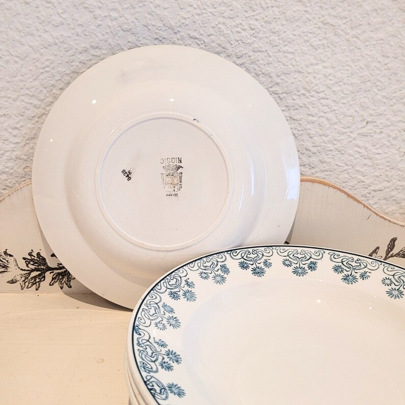 Może przedstawiać: Zestaw białych ceramicznych talerzy z niebieskim wzorem kwiatowym wok&oacute;ł krawędzi. Tylna strona jednego talerza zawiera znak producenta "DIGOIN."