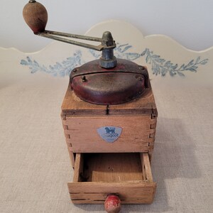May include: Vintage wooden coffee grinder with a red and metal top. The grinder has a drawer and a wooden handle. The Peugeot logo is visible on the front. The grinder is likely from the early to mid-20th century.