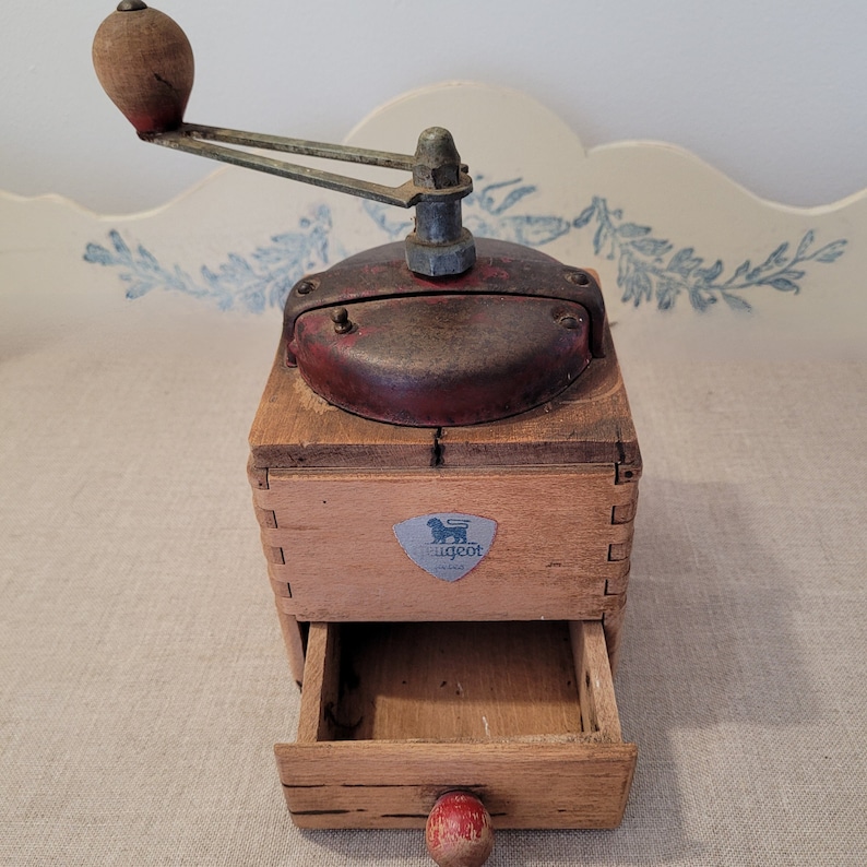 May include: Vintage wooden coffee grinder with a red and metal top. The grinder has a drawer and a wooden handle. The Peugeot logo is visible on the front. The grinder is likely from the early to mid-20th century.