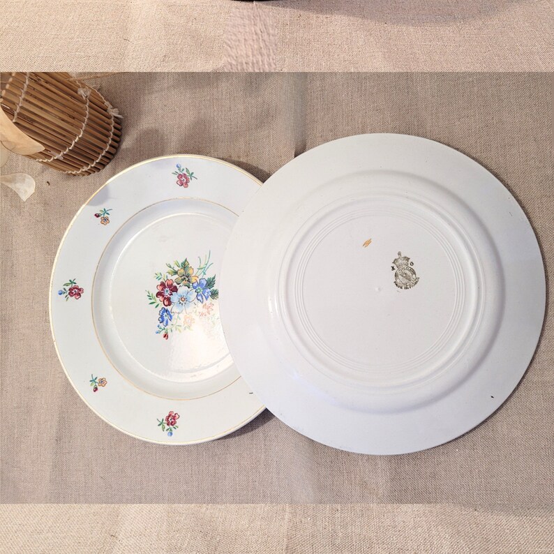 May include: Two white ceramic plates with a gold rim. The top plate has a floral design with pink, blue, and yellow flowers. The bottom plate is plain white.