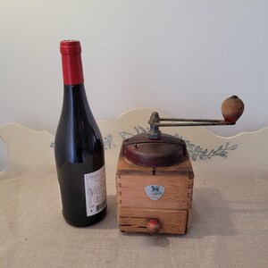 May include: An antique wooden Peugeot coffee grinder with a red handle and a small drawer, next to a dark wine bottle with a red cap. The grinder has a metal top and a Peugeot logo.