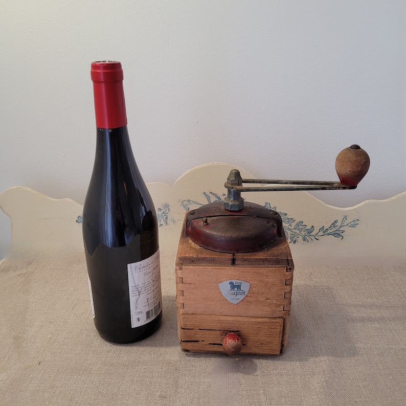 May include: An antique wooden Peugeot coffee grinder with a red handle and a small drawer, next to a dark wine bottle with a red cap. The grinder has a metal top and a Peugeot logo.