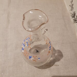 K&ouml;nnte beinhalten: Eine klare Glasflasche mit einer weiten &Ouml;ffnung und einem Blumendesign. Die Flasche hat einen rosa und wei&szlig;en Hintergrund mit braunen und blauen floralen Akzenten.