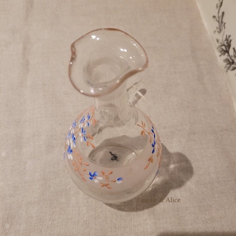 K&ouml;nnte beinhalten: Eine klare Glasflasche mit einer weiten &Ouml;ffnung und einem Blumendesign. Die Flasche hat einen rosa und wei&szlig;en Hintergrund mit braunen und blauen floralen Akzenten.