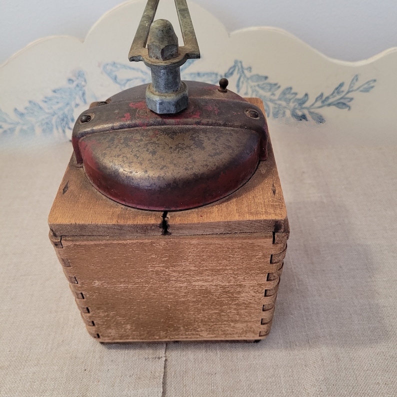 May include: An antique coffee grinder with a red and silver metal top and a wooden box base. The grinder has a crank handle and a small knob on top. The wood box is constructed with dovetail joints.