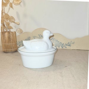 May include: White ceramic dish with a duck-shaped lid. The dish has a black rim and the duck has a slightly open beak.