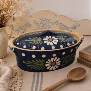 May include: An oval ceramic casserole dish with a deep blue glaze, featuring hand-painted white daisy motifs with orange centres and green leaf accents. The dish includes a matching lid with a central handle and a light tan, unglazed rim edge.