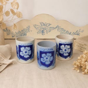 May include: Three cylindrical ceramic cups featuring a white base with a blue shield design. Each cup displays a hand-painted blue flower inside the shield and the word "Haguenau" written in blue script above the emblem. The cups have a smooth finish.