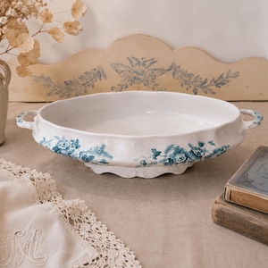 May include: An oval white ceramic serving dish with two side handles, featuring a blue floral transferware pattern along the exterior rim. The dish has a scalloped edge and a raised base, presenting a classic design for serving food at the table.
