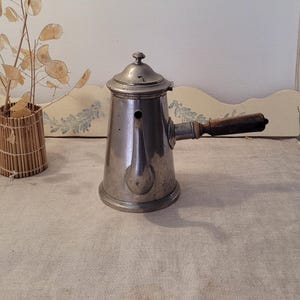 Può includere: Una caffettiera vintage color argento con manico in legno. La caffettiera ha un beccuccio e un coperchio con un piccolo pomello. Il manico è marrone scuro. La caffettiera è su una superficie beige.