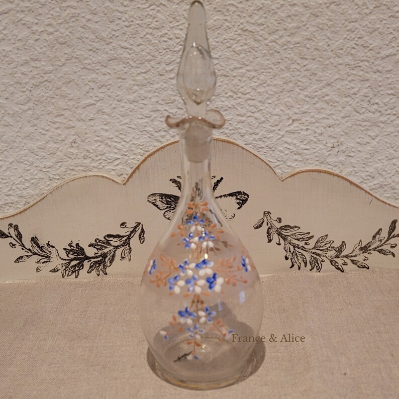 K&ouml;nnte beinhalten: Eine klare Glasflasche mit einem floralen Design in Blaut&ouml;nen, Wei&szlig; und Orange. Die Flasche hat einen langen, d&uuml;nnen Hals und einen Stopfen mit einem dekorativen Oberteil. Die Flasche steht auf einer wei&szlig;en Oberfl&auml;che mit einem dekorativen Design.