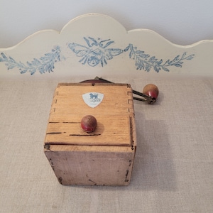 May include: Vintage wooden Peugeot coffee grinder with a crank handle and a red knob. The grinder has a rectangular shape with dovetail joints and a Peugeot logo. The wooden box is light brown with a red knob on top.
