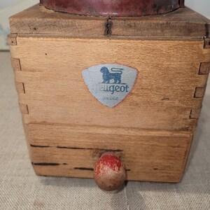 May include: A vintage wooden coffee grinder with a red metal top. The wooden body features dovetail joints and a small, red and white knob. A silver label with a lion logo and the word "Peugeot" is affixed to the front.