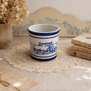 Peut inclure: Un pot en céramique blanc et bleu avec le texte "Bournemouth Old English" et une illustration de château. Le pot a un bord bleu et deux rayures bleues. Il repose sur un napperon en dentelle.