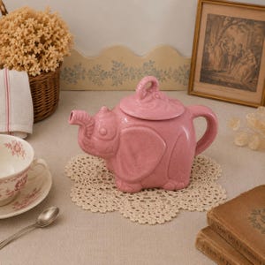 Puede incluir: Una tetera de cerámica rosa con forma de elefante, con tapa a juego. La tetera tiene una superficie texturizada y está sobre un tapete color crema. Una taza y un platillo están en primer plano.