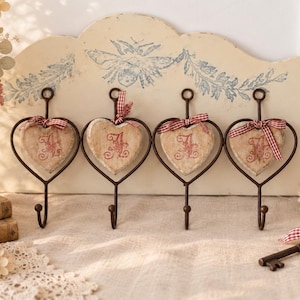 May include: A wall-mounted metal rack featuring four heart-shaped hooks. Each hook holds a distressed, cream-colored wooden heart ornament with a red embroidered letter A and a small red and white checkered ribbon tied at the top loop.