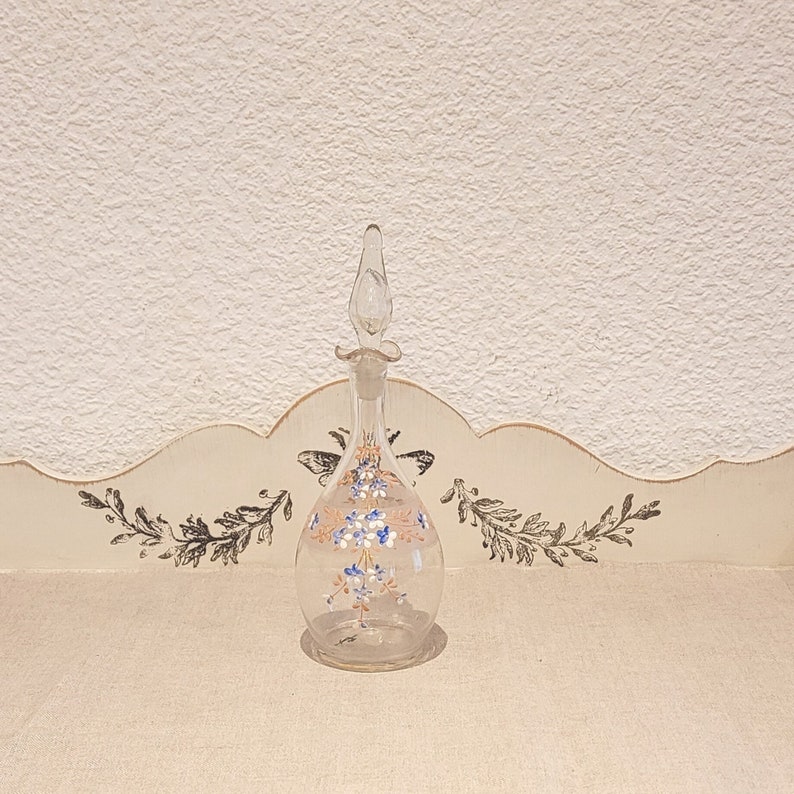 K&ouml;nnte beinhalten: Eine klare Glasflasche mit einem floralen Design in Blaut&ouml;nen, Wei&szlig; und Orange. Die Flasche hat einen Stopfen und einen runden K&ouml;rper.