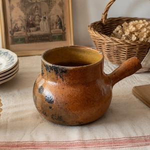 May include: A rustic, brown ceramic pot with a handle. The pot has a rounded body and a wide opening, with dark spots and a yellow interior. The pot is sitting on a cream-coloured tablecloth.
