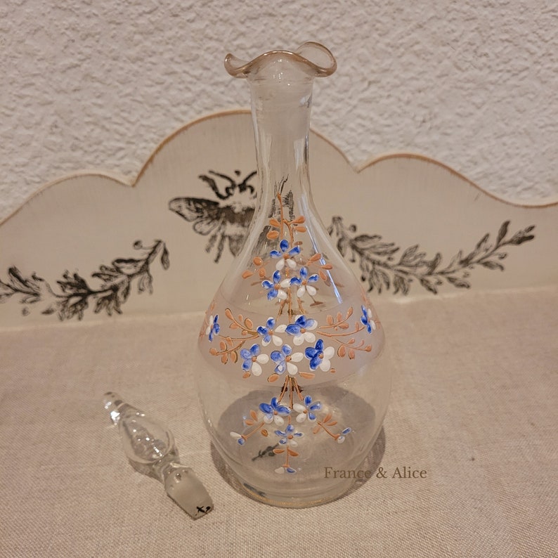 K&ouml;nnte beinhalten: Eine klare Glasflasche mit einem floralen Design, das in Blau, Wei&szlig; und Orange bemalt ist. Die Flasche hat einen ausgestellten oberen Teil und einen Stopfen.