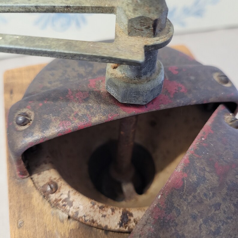 May include: Close-up of an antique coffee grinder. The grinder features a red and black metal body with a wooden base. The handle and other metal components show signs of age and wear. The grinder is designed to grind coffee beans.