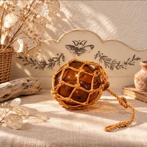 May include: A spherical amber glass float encased in a thick, knotted tan jute rope netting. The glass orb features a smooth, reflective surface, while the attached rope includes a sturdy loop at one end, designed for hanging or decorative display purposes.