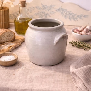Pode incluir: Um pote de cerâmica cinza claro com duas alças, sobre uma toalha de mesa de linho de tom neutro. O pote é cercado por ingredientes como pão, azeite, alho e sal, sugerindo um tema culinário.