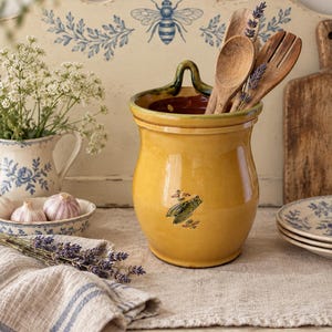 May include: A yellow ceramic utensil crock with a glossy finish and a small green cicada illustration on the front. The container features a curved top edge with a central handle loop and holds several wooden kitchen spoons and a sprig of lavender.