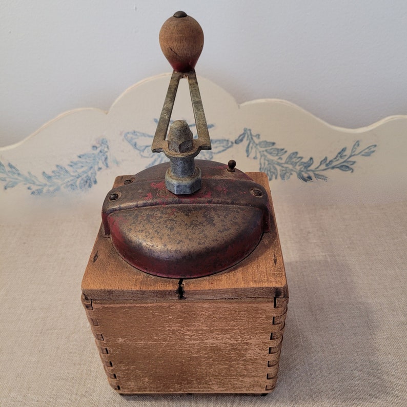 May include: An antique coffee grinder with a wooden base and a red and metal top. The grinder features a wooden handle and a metal crank. The wooden base is constructed with dovetail joints. The grinder is likely from the early 20th century.