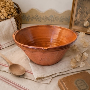 May include: A large, terracotta mixing bowl with a wide rim and two small handles. The bowl has a glossy finish and a warm, earthy tone. The text "Foterik Nice" is visible on the side. A wooden spoon and linen cloth are in the foreground.