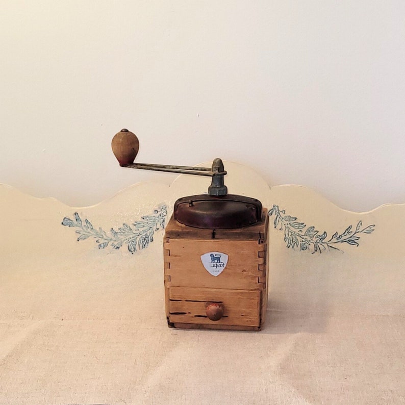 May include: A vintage wooden coffee grinder with a metal crank and a small drawer. The grinder has a wooden handle and a metal top. The Peugeot logo is visible on the front. The grinder is set on a white surface.