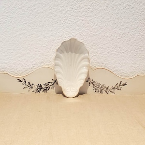 May include: A white ceramic seashell-shaped dish with a raised design on the bottom. The dish is sitting on a white surface with a decorative border.