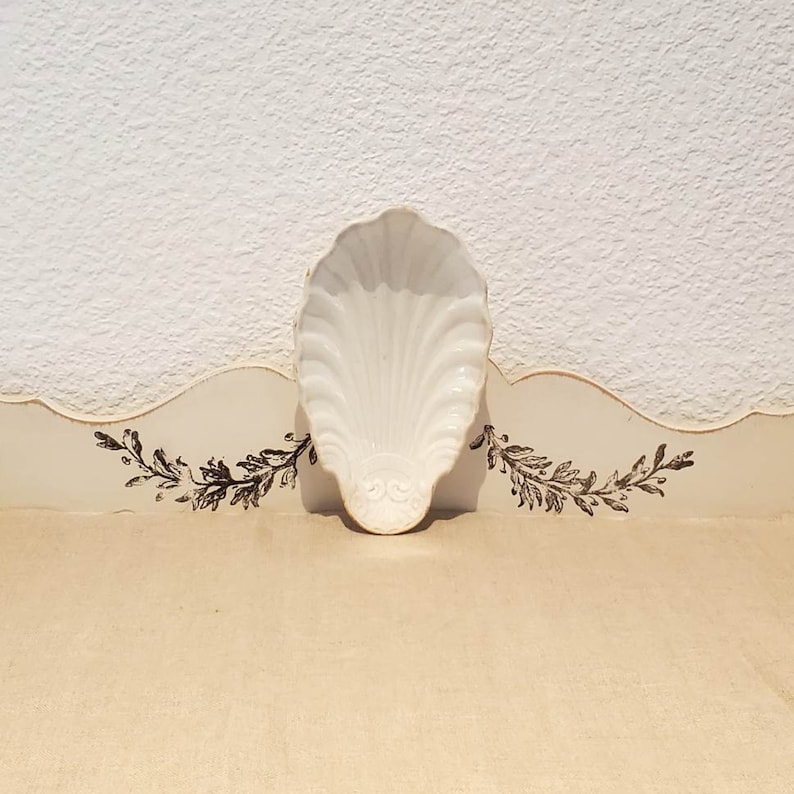 Puede incluir: Un plato de cer&aacute;mica blanca con forma de concha marina con un dise&ntilde;o en relieve en la parte inferior. El plato est&aacute; sobre una superficie blanca con un borde decorativo.