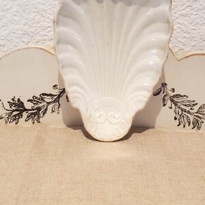 Puede incluir: Plato de cer&aacute;mica blanca en forma de concha con un dise&ntilde;o decorativo. El plato est&aacute; sobre una superficie blanca con un patr&oacute;n floral negro.