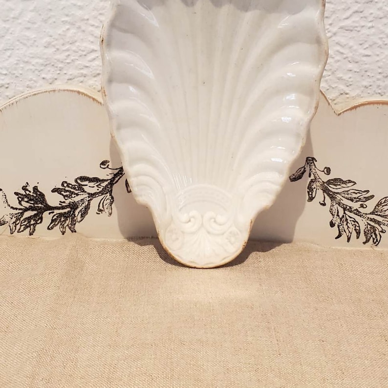 Puede incluir: Plato de cer&aacute;mica blanca en forma de concha con un dise&ntilde;o decorativo. El plato est&aacute; sobre una superficie blanca con un patr&oacute;n floral negro.