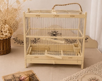 Op de afbeelding: Een rechthoekige, crèmekleurige vogelkooi met een draadgaasstructuur, een touwgreep en een zwarte bijengrafiek op het achterpaneel. De kooi is voorzien van een kleine houten zitstok, een voerbakje en decoratieve bloemmotieven op het frame.