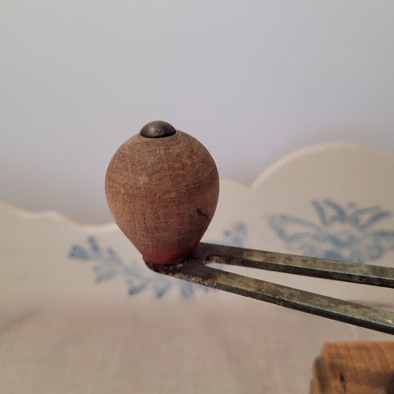 May include: Close-up of a vintage wooden kitchen tool. The tool features a rounded, brown wooden handle with a small, dark metal cap. The handle is attached to a metal piece with a fork-like design. The tool rests against a white surface with blue floral patterns.