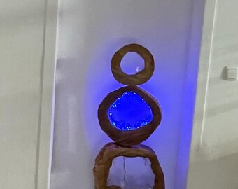 Puede incluir: Una escultura de madera única con un diseño apilado. La escultura presenta tres formas abiertas e irregulares, con una luz azul que ilumina el centro de la forma central. La escultura está colocada sobre una base gris oscuro.