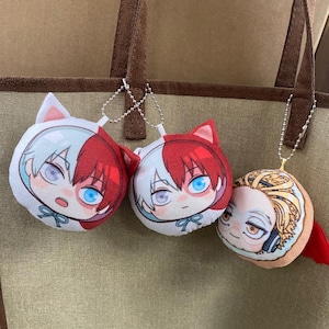May include: Three plush anime character keychains with white, red, and yellow faces. The characters have white, red, and blonde hair. The keychains are attached to brown tote bags.